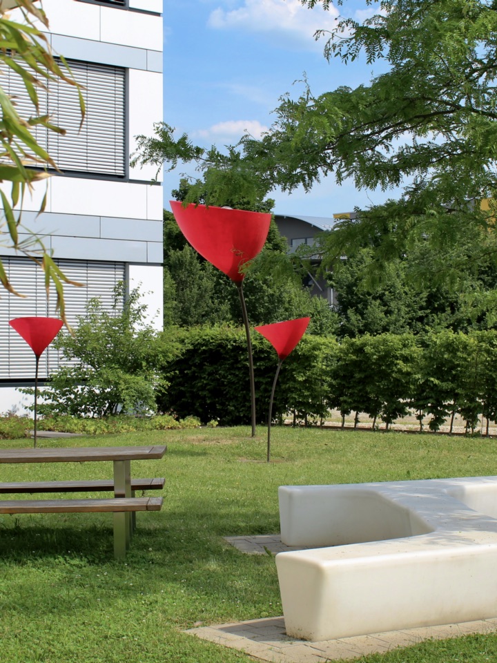 In the garden of the MVC, a classic bench and a white seat in the shape of an "I" can be seen. A small tree and some bushes are in the background, while red lamps that look like poppies illuminate the picture.