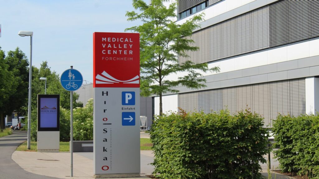 Deutlich erkennbar ist das Schild des Medical Valley Forchheim, das in Rot mit weißer Schrift oben angebracht ist. Der untere Teil des Schildes ist grau, und der Name "HiroSakao" ist daneben zu sehen, begleitet von einem Hinweis zur Einfahrt zum Parkplatz.