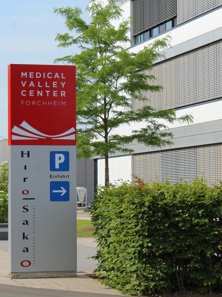 The Medical Valley Forchheim sign is clearly visible, in red with white lettering at the top. The lower part of the sign is grey, and the name "HiroSakao" can be seen next to it, accompanied by a sign indicating the entrance to the car park.