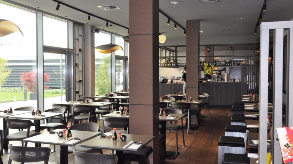 The interior of Hiro Sakao is decorated in a dark grey and brown, with a modern ambience. Tables with 2 chairs each are spread out, while two posts decorate the space and the kitchen can be seen in the background.
